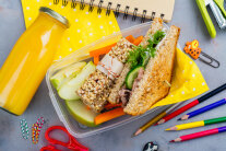 Zwischen Stiften und Bastelmaterial steht ein Sandwich mit Gemüse und Obst in einer Box, daneben liegt eine Flasche mit Orangensaft
