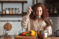 Junge Frau steht in einer modernen Küche und kostet Essen aus einem Schöpflöffel