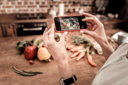 Eine Frau fotografiert mit einem Smartphone einen rustikalen Küchentisch mit Lebensmitteln