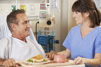 Mann im Klinikbett nimmt lächelnd Tablett mit Essen von Pflegefachkraft entgegen